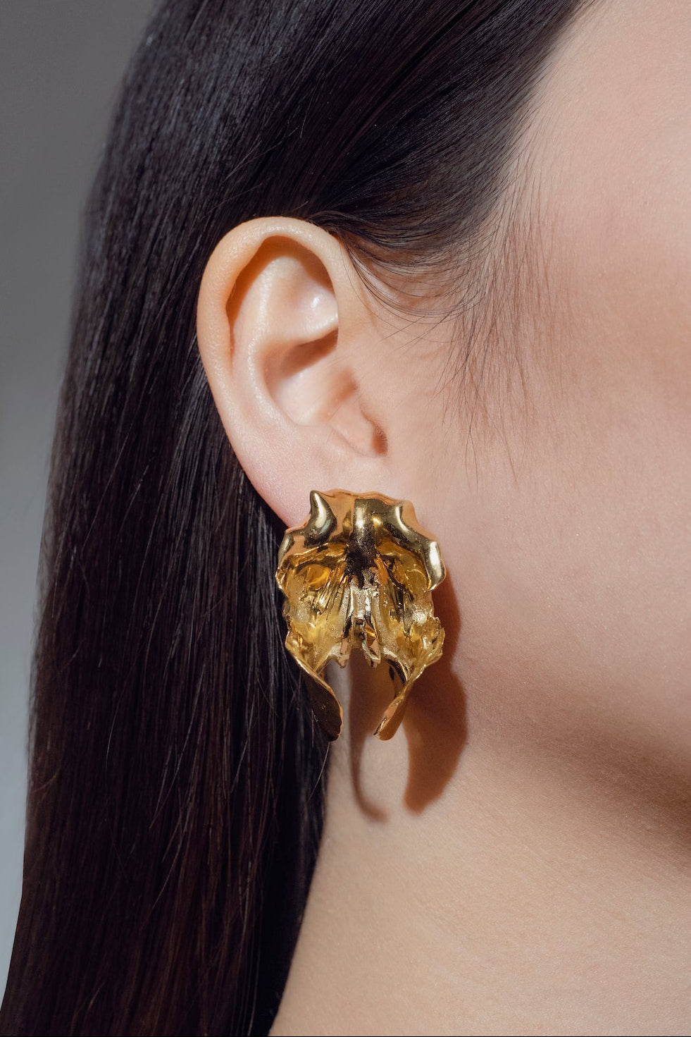 Gold orchid-shaped earring worn by a model with dark hair on agrey background