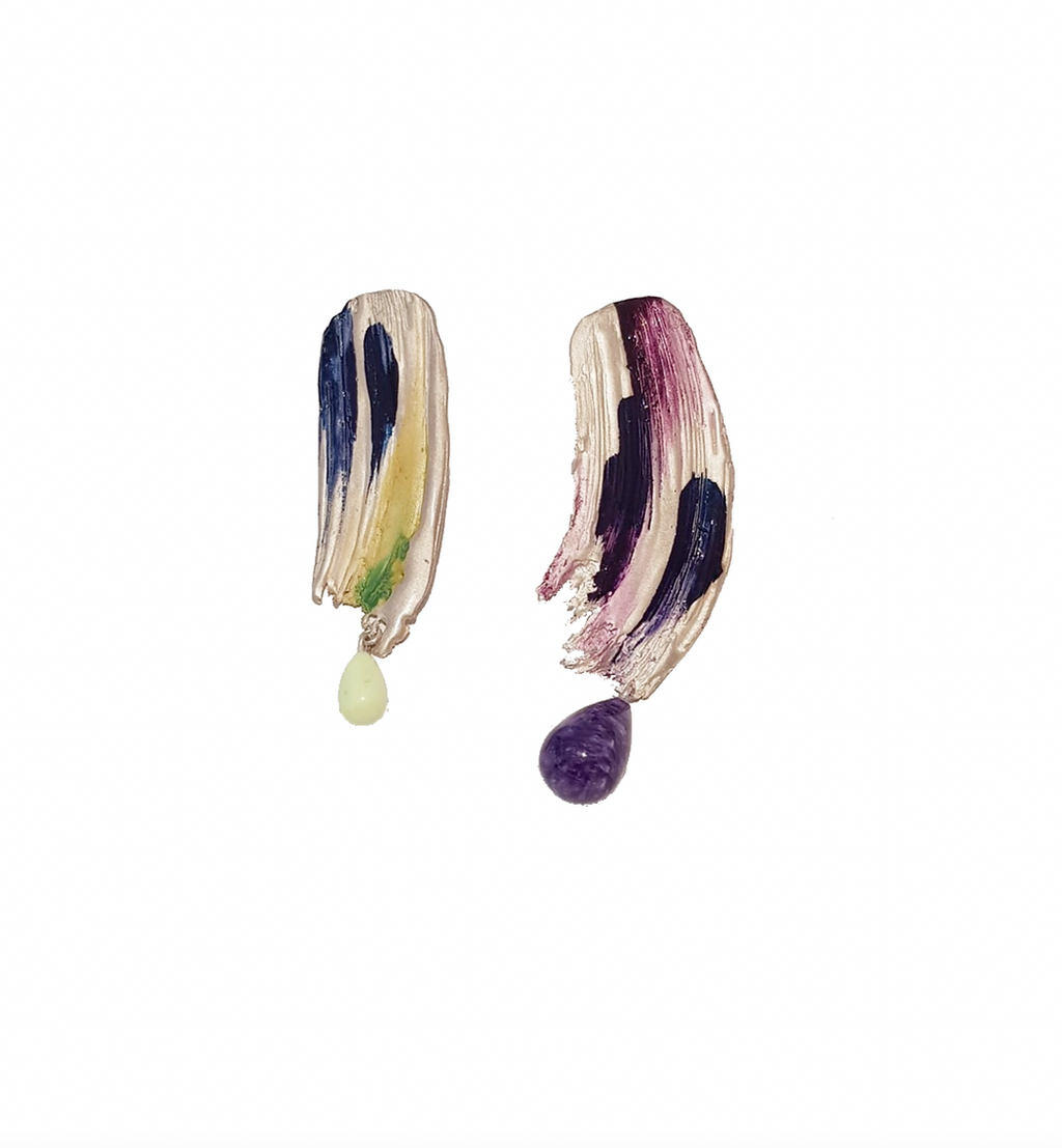 Handcrafted earring with blue, purple, yellow and a bit of green painted on it with two different gemstones, on the left a white one and on the right a purple gemstones. they are displayed on awhite background