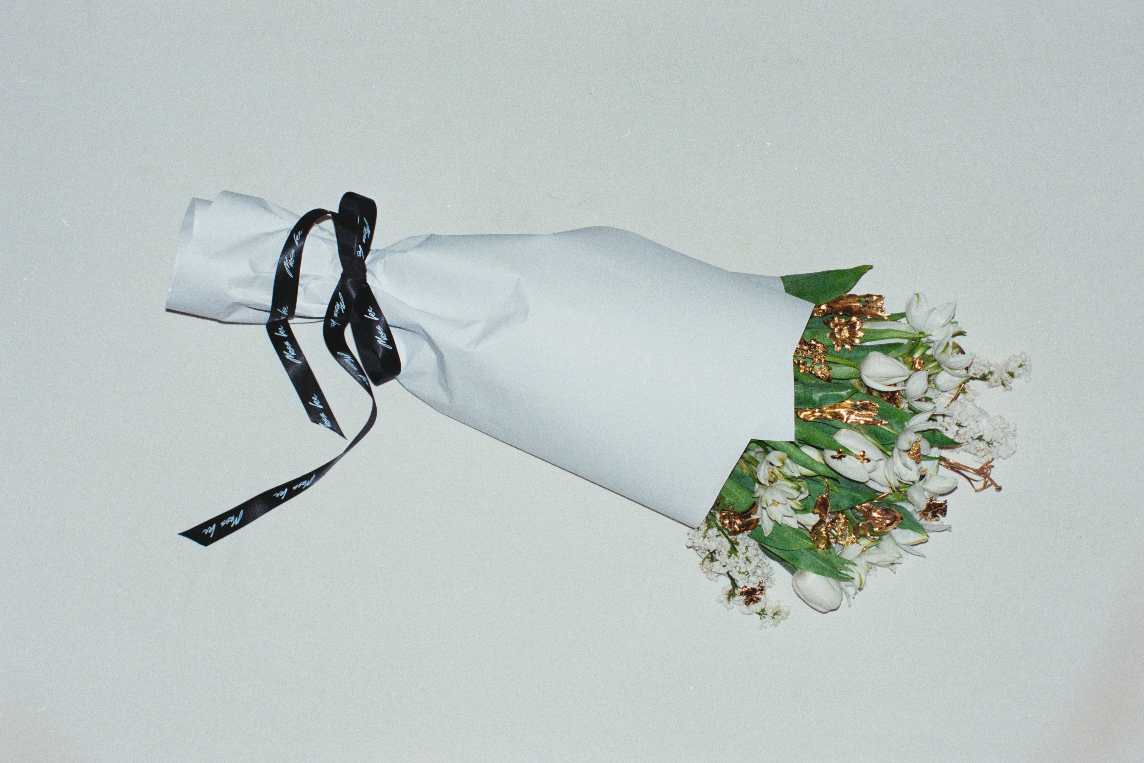 Bouquet of flowers with handcrafted gold jewlery in between, wrapped in white paper with a black ribbon on a light gray background