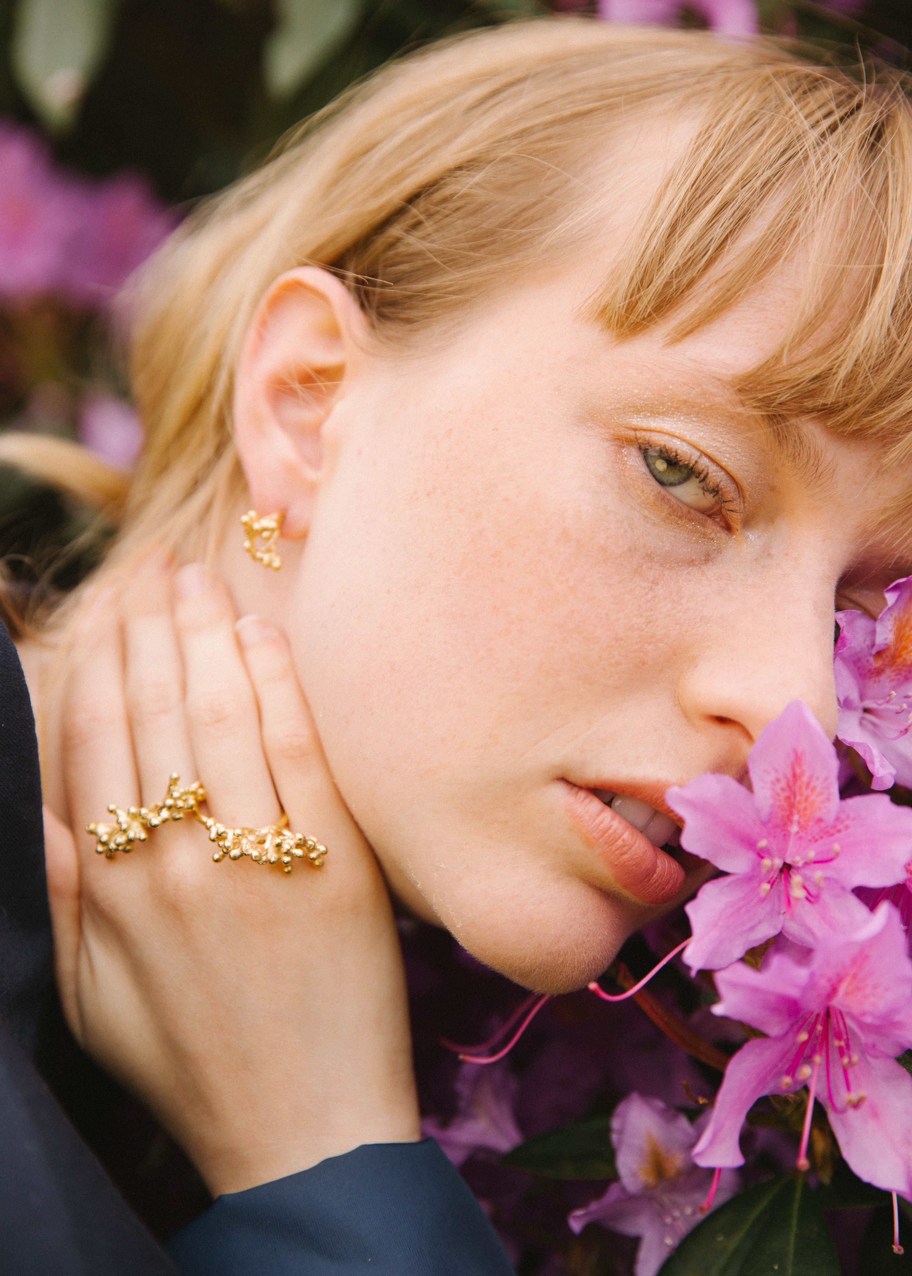 Woman wearing handcrafted gold earrings and rings with pink flowers in the background