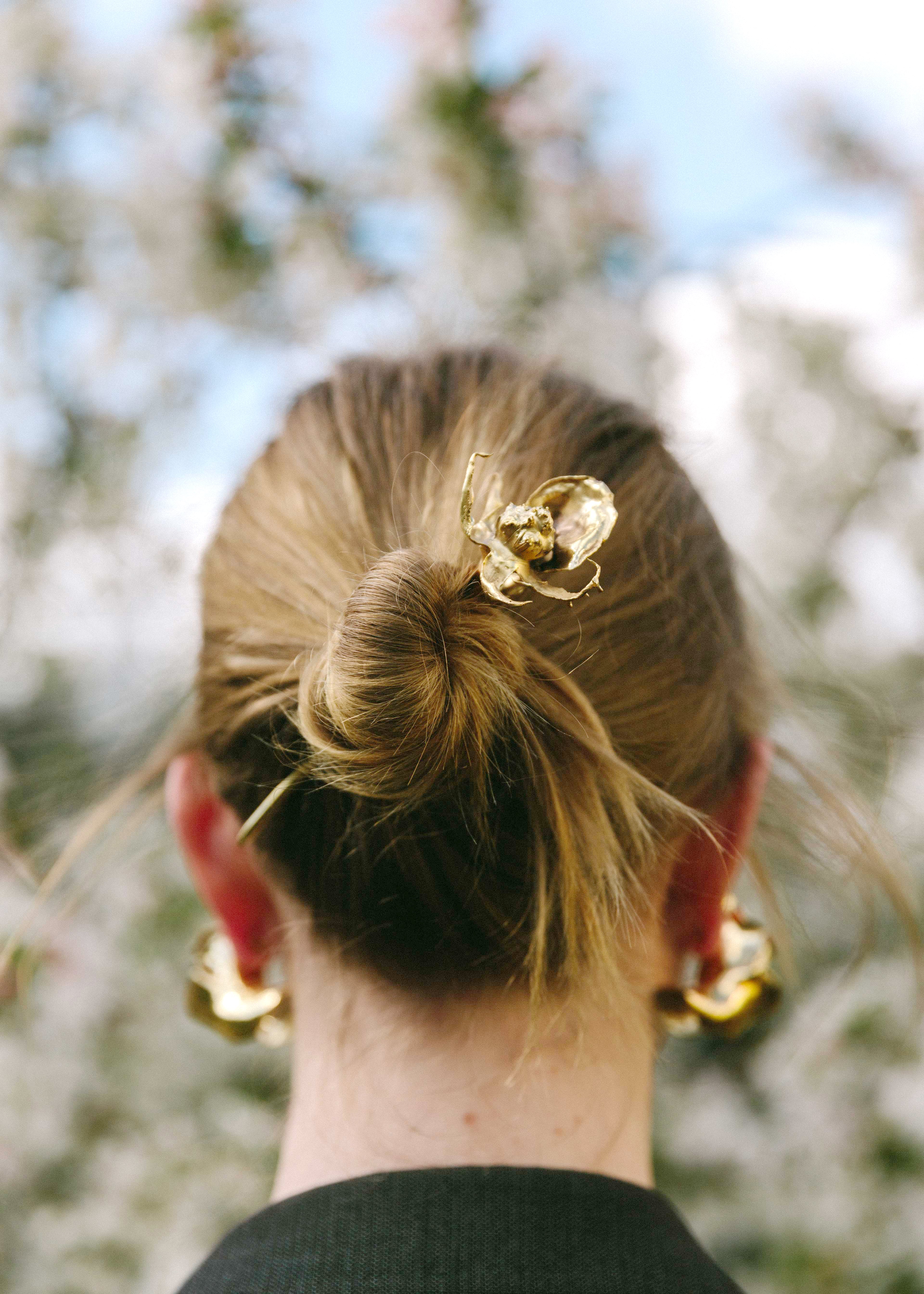 Person with a bun hairstyle featuring a handcrafted decorative rose hair clip, wearing gold earrings, with a blurred natural background.